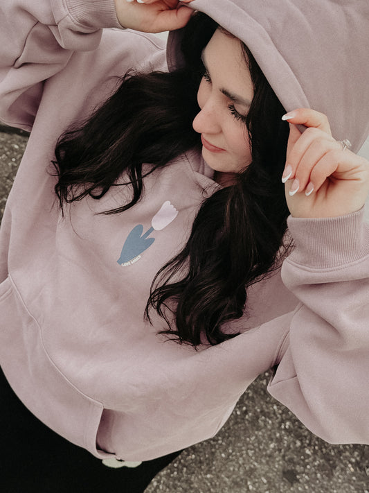 Model wearing LOVE WORLD Manifest Hoodie in Dark Purple — close-up of oversized dusty mauve fleece hoodie with tulip chest graphic, pulling up hood, outdoor shot