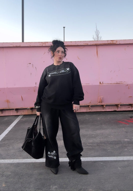 Model wearing LOVE WORLD MOM WORLD Monkey Wash Sweatshirt and jogger set in black — back view showing Mom World star graphic, styled with heeled boots against pink wall