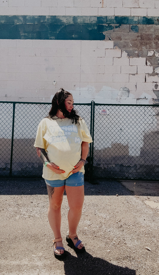 Pregnant mom wearing the LOVE WORLD Happy As A Mother Tee in Banana — oversized yellow garment-dyed tee with retro bubble letter graphic, styled with denim shorts and platform sandals