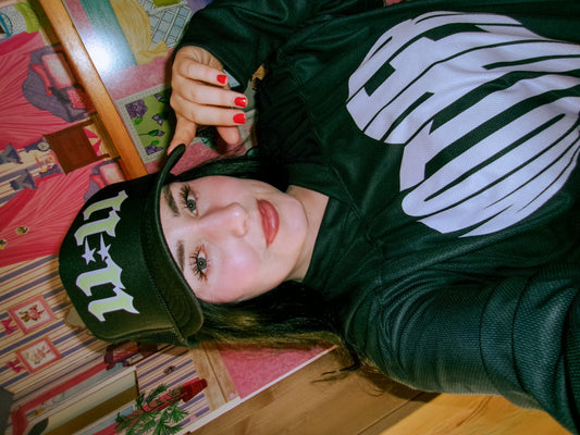 Woman wearing the LOVE WORLD 11:11 Trucker Hat in black with neon green gothic lettering — styled with MOTHER graphic hoodie, playful dollhouse backdrop