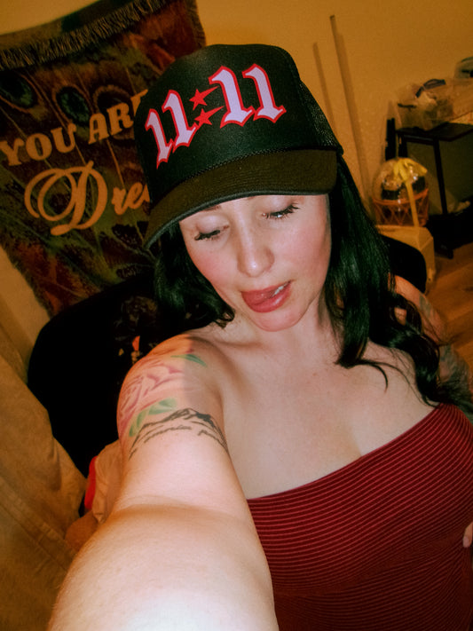 Woman wearing the LOVE WORLD 11:11 Trucker Hat 2.0 in black — bold red and pink gothic embroidery, selfie shot