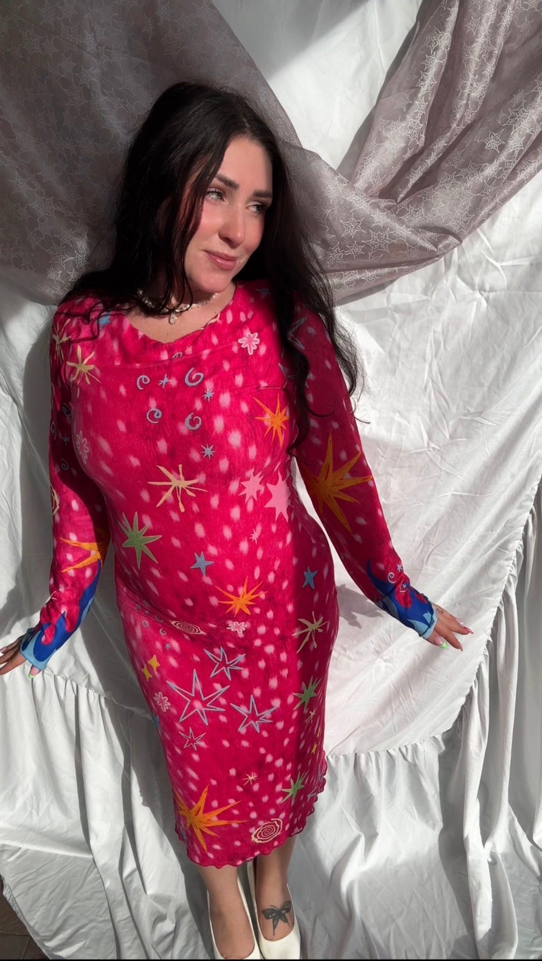 Model wearing LOVE WORLD’s hot pink Deer Stars mesh midi dress with colorful stars, spirals and blue flame cuffs against draped white fabric.