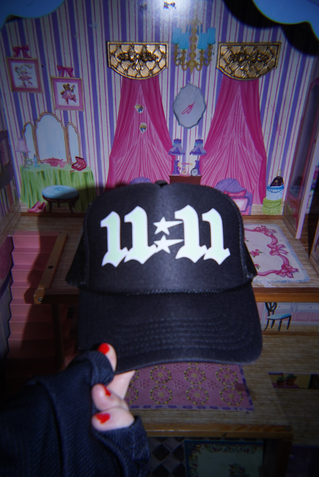 Hand holding the LOVE WORLD 11:11 Trucker Hat facing forward — black foam front with bold white gothic 11:11 embroidery and star, whimsical dollhouse background