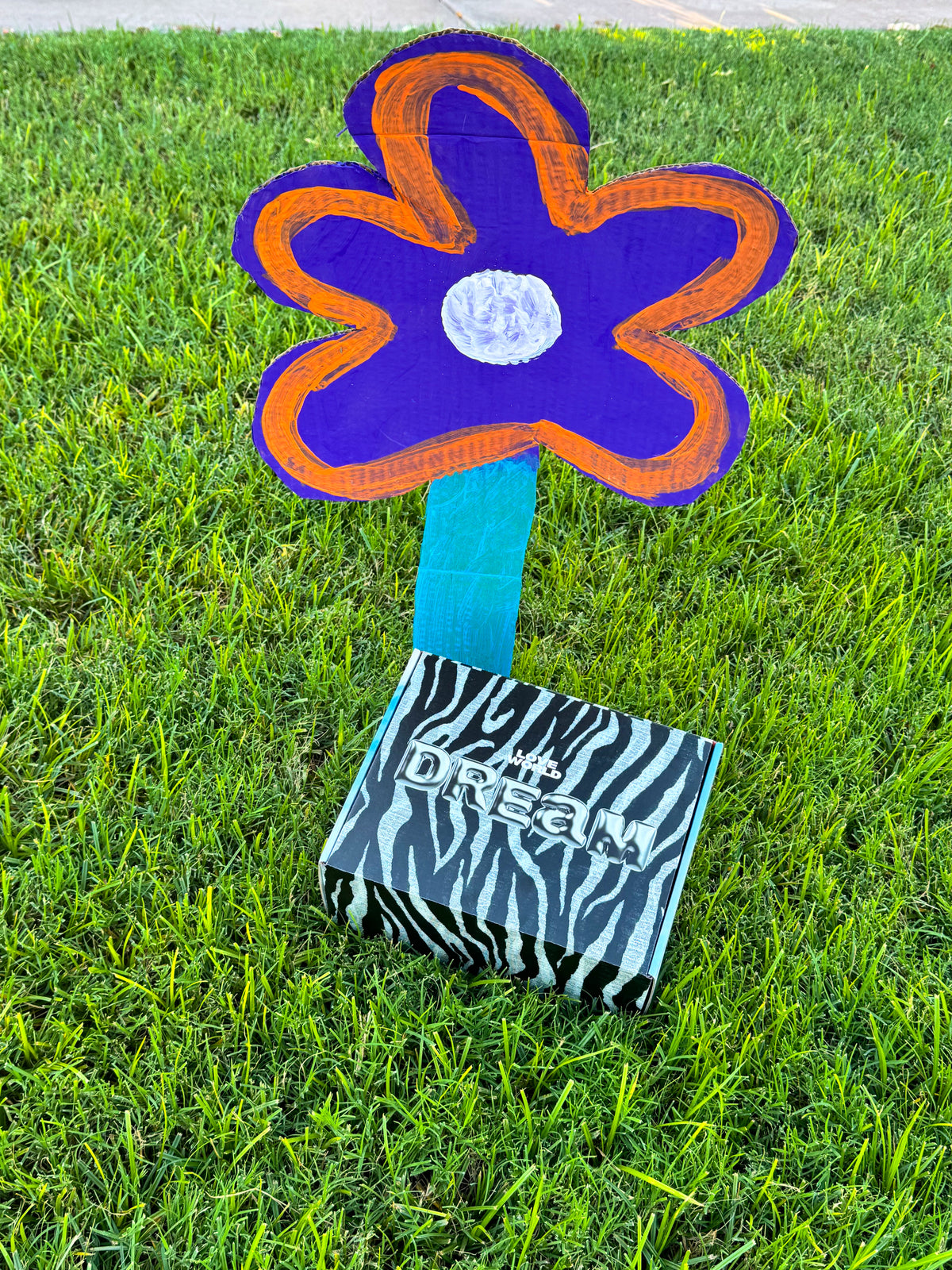 A handmade flower yard sign with a purple and orange painted flower head, a turquoise stem, and a zebra-print box base printed with the word 'DREAM', placed on green grass outdoors.