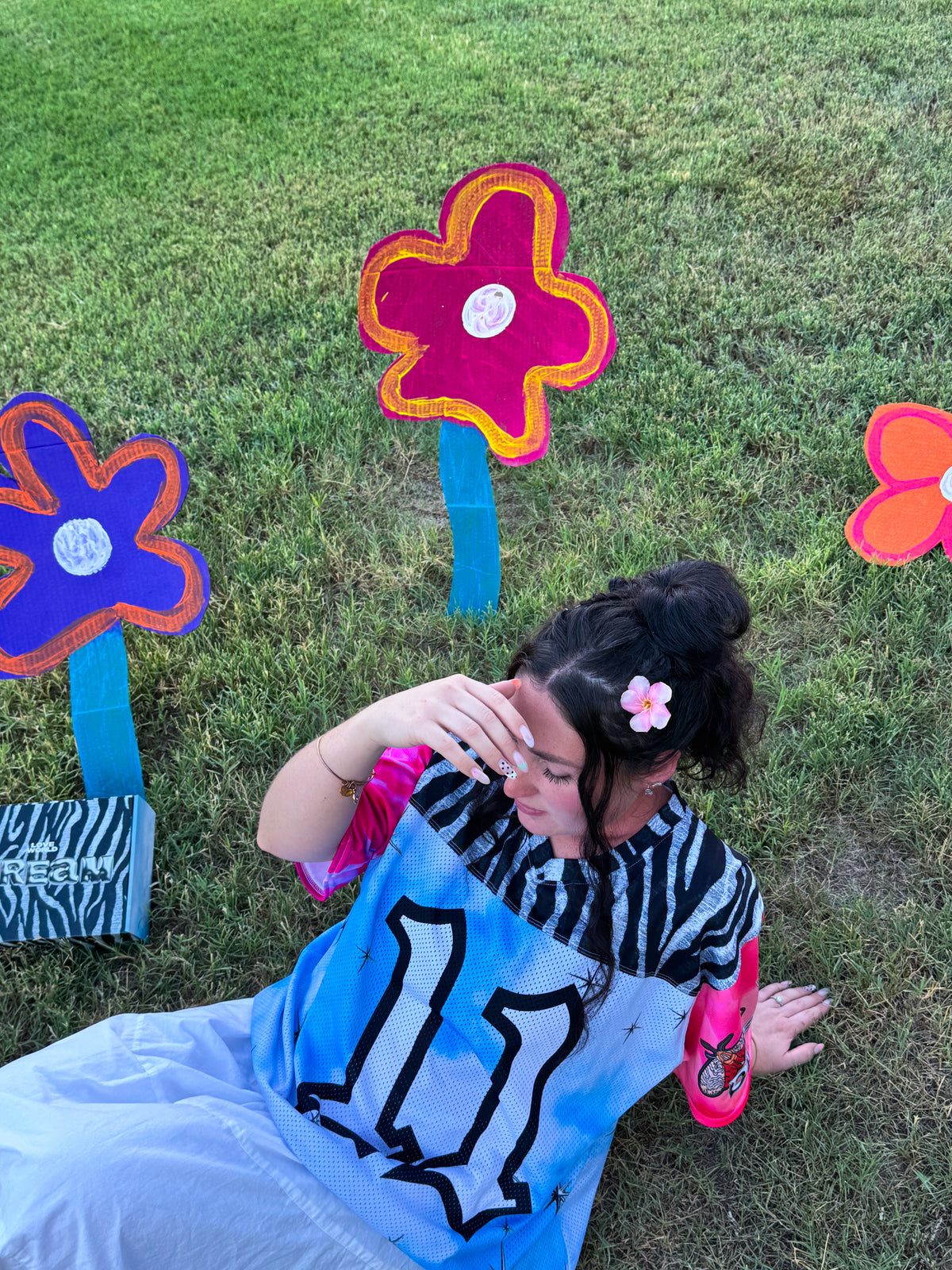 A person sitting on grass outdoors wearing a light blue oversized jersey with a black number 11, a black-and-white striped top, and a pink flower hair clip. Colorful flower-shaped signs are placed around them on the lawn.