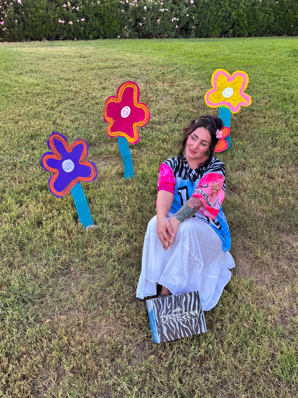 A woman sits on a grassy lawn wearing a colorful outfit with a white skirt, next to three large flower-shaped garden signs. A zebra-print box labeled 'GREATLY DESIGNED' rests on the grass in front of her.