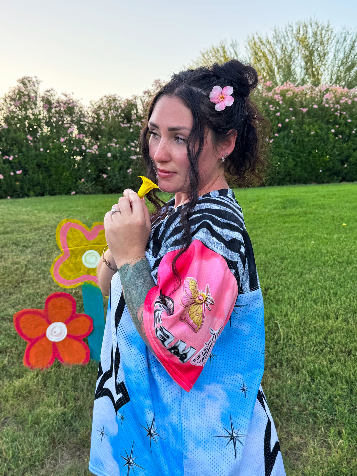 A woman standing outdoors on a grassy lawn wearing a colorful, patterned poncho-style garment. The garment features a black and white zebra stripe pattern on the upper section, a bright pink section with a bee graphic and text, and a light blue lower section with black star-like patterns.