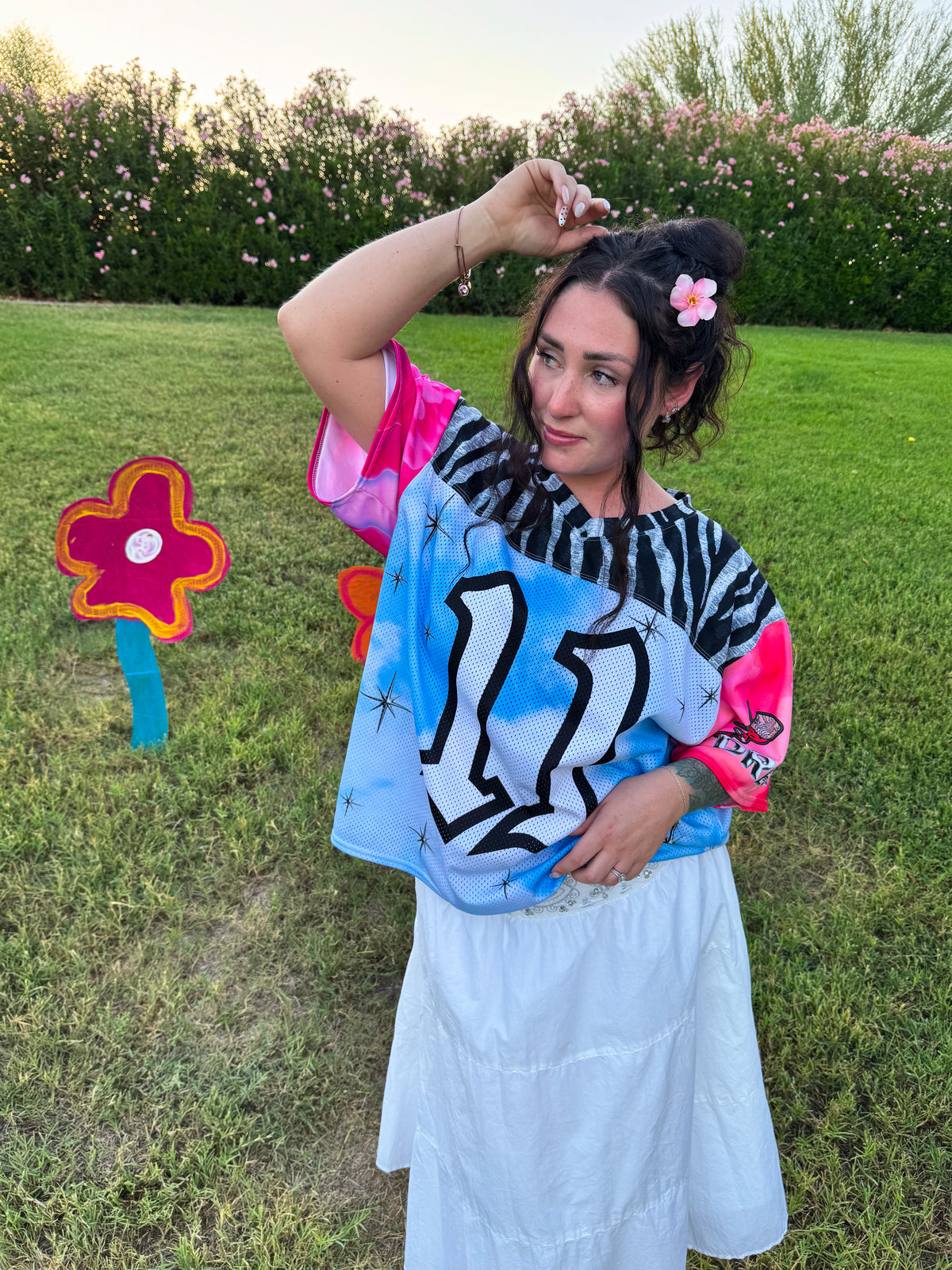 A woman stands outdoors on a grassy lawn wearing a colorful graphic t-shirt with pink sleeves, a blue and black patterned body, and a white skirt. She has a pink flower in her hair, with flower-shaped yard signs on the grass to her side.