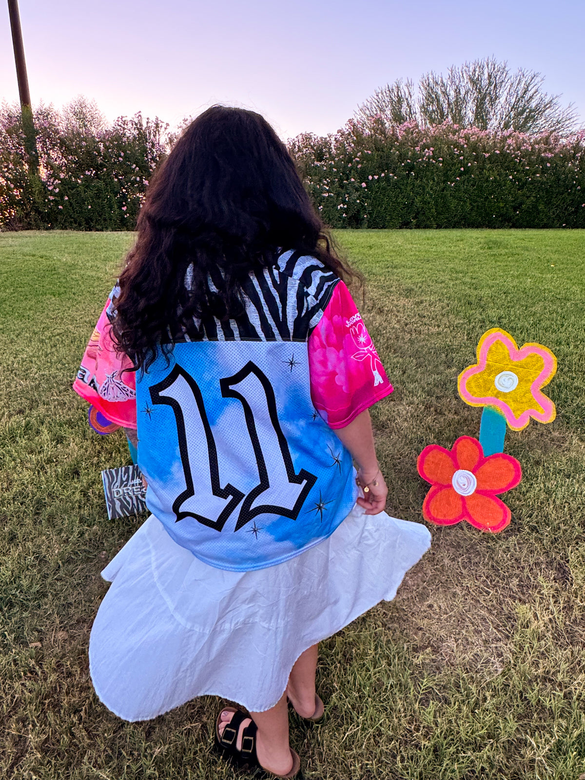 A person seen from behind wearing a colorful graphic jersey with the number 11, standing on a grassy lawn outdoors. The jersey has a light blue back with a large black outlined number 11, bright pink sleeves, and black-and-white zebra print shoulder panels.