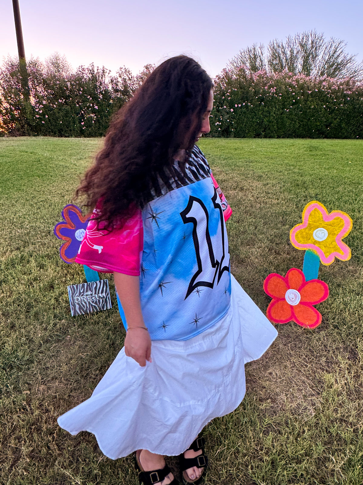 A woman outdoors on a grassy lawn at sunset, wearing a colorful t-shirt with a large black number 11 on a light blue background, a white skirt, and black sandals. She is holding a zebra-print bag, with decorative flower yard signs nearby.