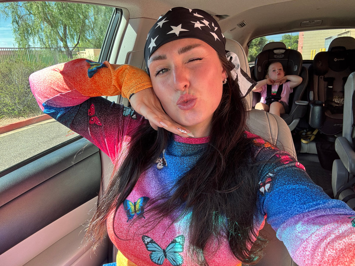 A woman taking a selfie inside a car, wearing a black bandana with white stars and a long-sleeve colorful top with a blue, pink, and orange gradient and butterfly print. A child is seated in a car seat in the background.