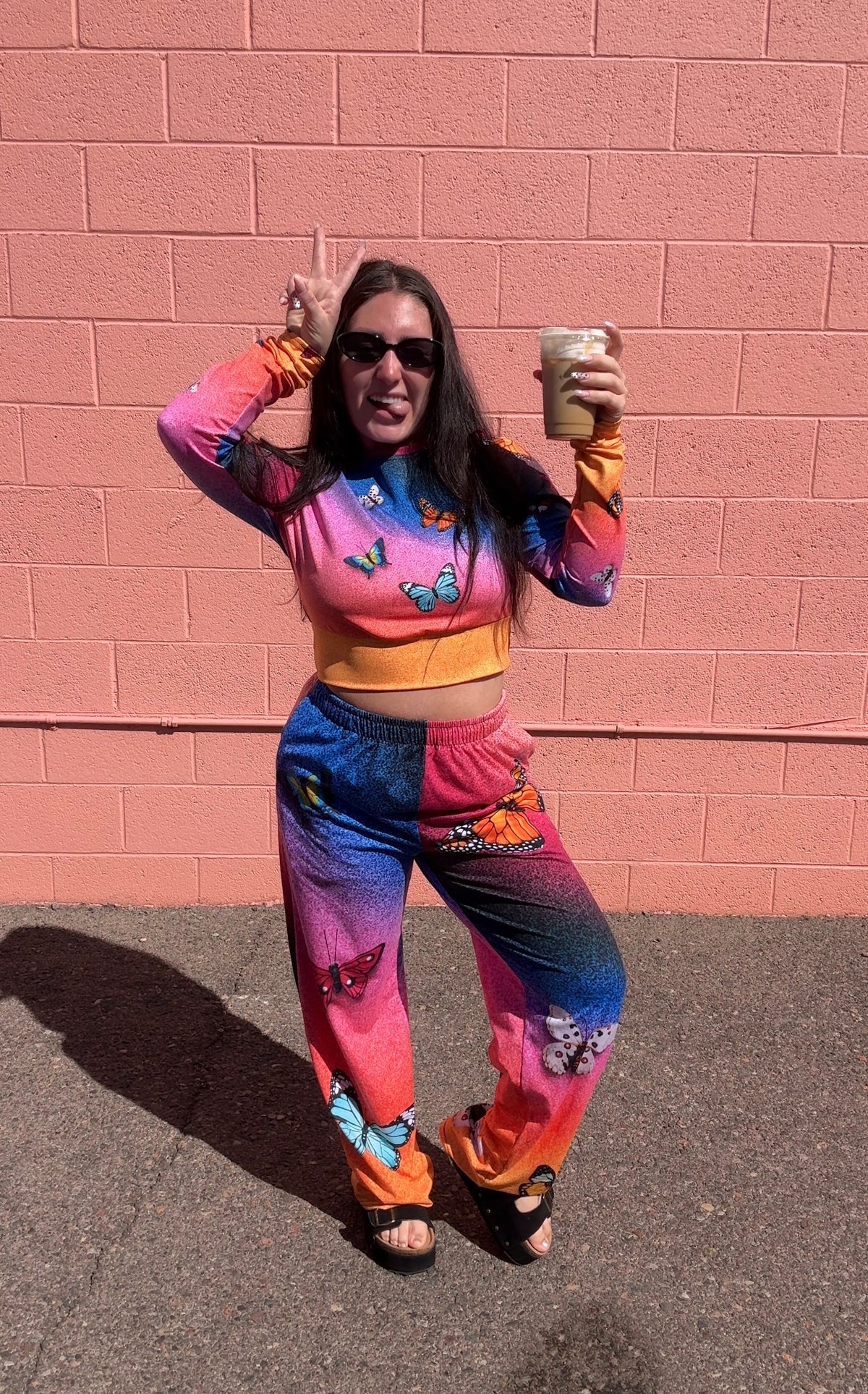 A woman posing outdoors in front of a pink brick wall, wearing a vibrant, multicolor long-sleeve crop top and matching sweatpants with butterfly prints and gradient patterns in pink, blue, orange, and purple.