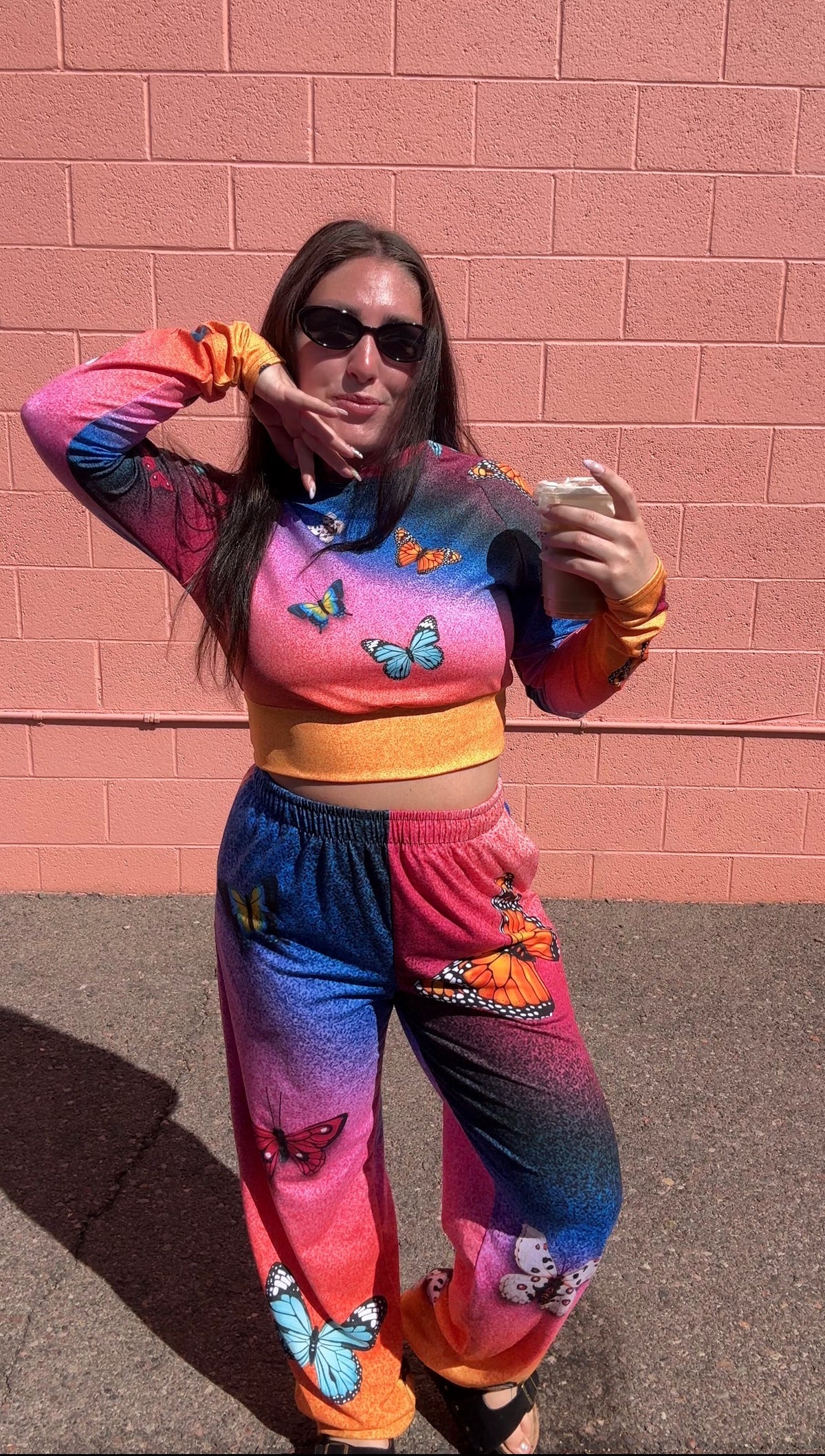 A woman wearing a matching two-piece lounge set with a vibrant rainbow gradient and butterfly prints, posing in front of a pink brick wall.
