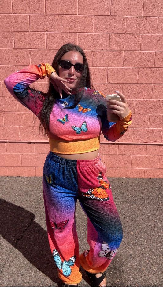 A woman wearing a matching two-piece lounge set with a vibrant rainbow gradient and butterfly prints, posing in front of a pink brick wall.