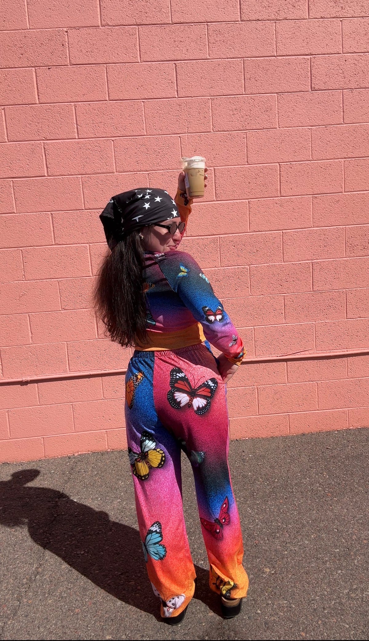 A person stands outdoors against a pink brick wall wearing a bright multicolor tie-dye outfit with butterfly prints, a black bandana, and holding a takeaway coffee cup.