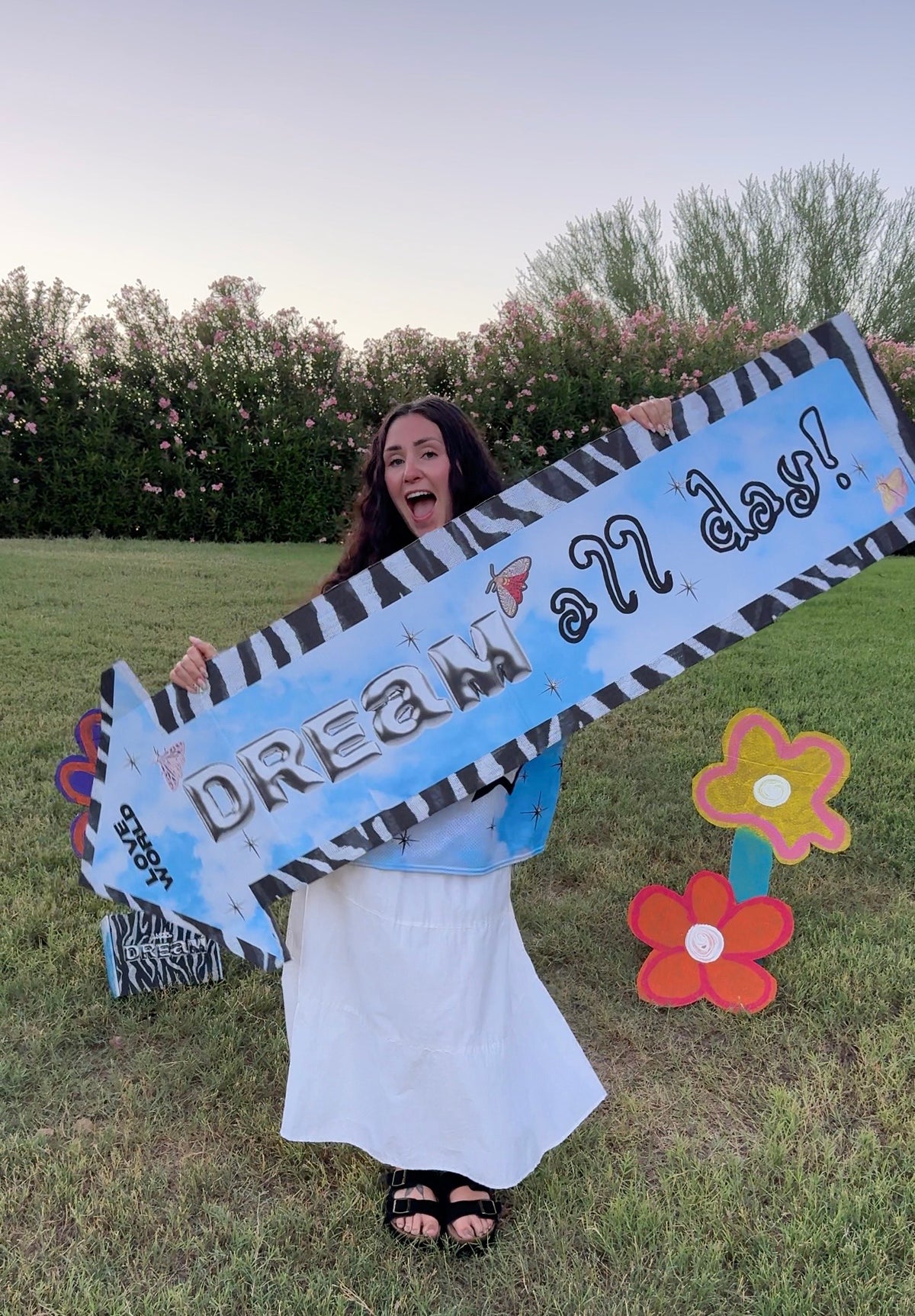 A woman stands outdoors on a grassy lawn holding a large decorative banner that reads 'DREAM all day!' with a blue and black zebra-stripe design. She is smiling broadly, with trees and bushes in the background and two colorful flower cutouts on the grass nearby.