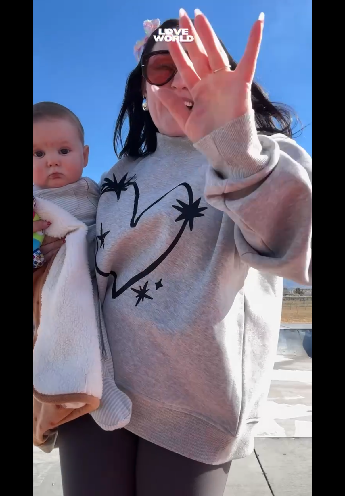 Mom holding a baby while wearing the LOVE WORLD Stardust Mock Neck Sweater in Flower Gray — hand-drawn heart and stardust graphic, relaxed oversized fit outdoors