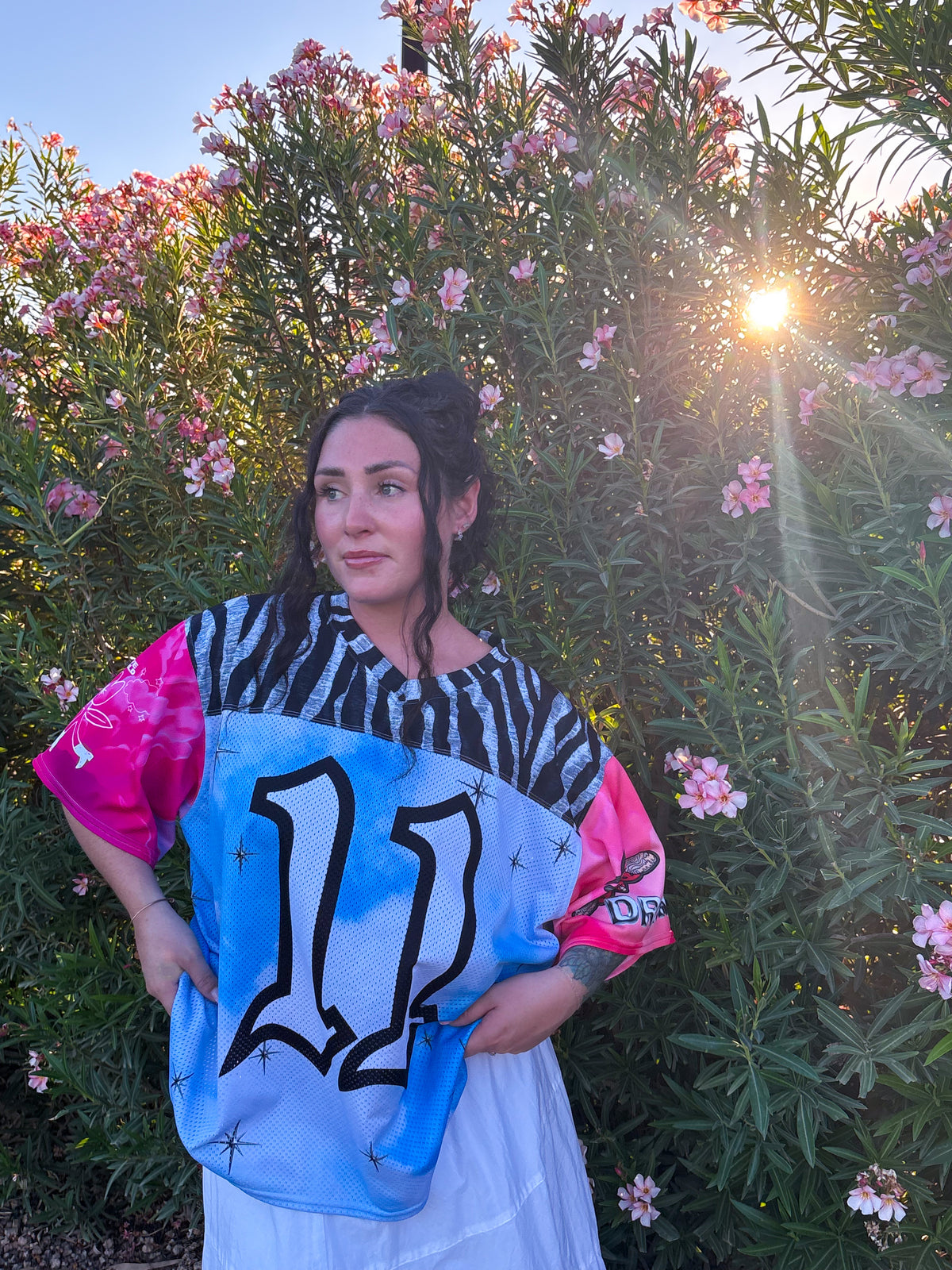 A woman stands outdoors in front of flowering bushes at sunset, wearing a colorful graphic T-shirt with a black-and-white zebra print upper section, bright pink sleeves, a light blue lower section, and a large black number 11 on the front.