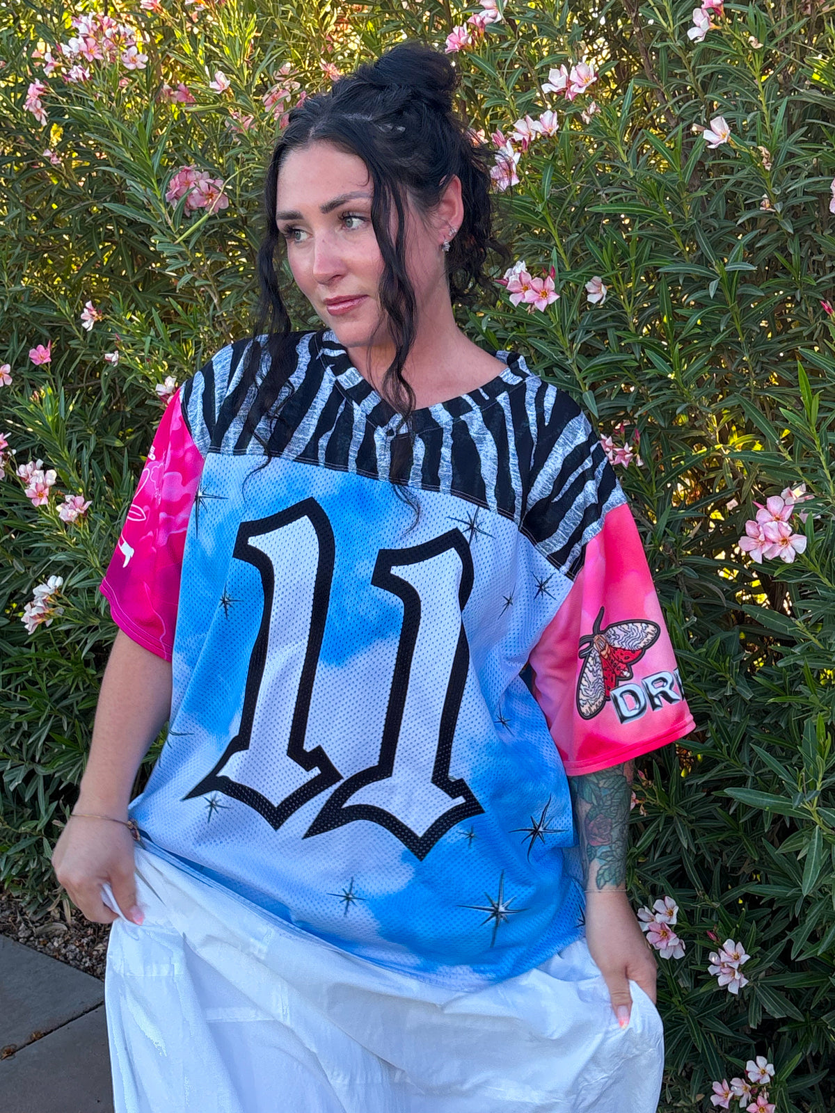 A woman wearing a colorful graphic t-shirt with a blue and white mesh front, black and white zebra print on the upper back, and bright pink sleeves, posing outdoors in front of green bushes with pink flowers.