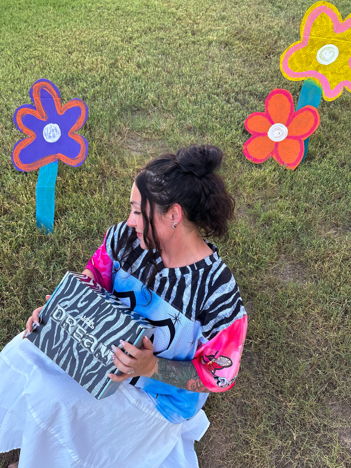 A woman sitting on a grassy lawn holds a black and white zebra-print box with the text 'DREAM'. She is wearing a colorful, patterned long-sleeve top and a white skirt.
