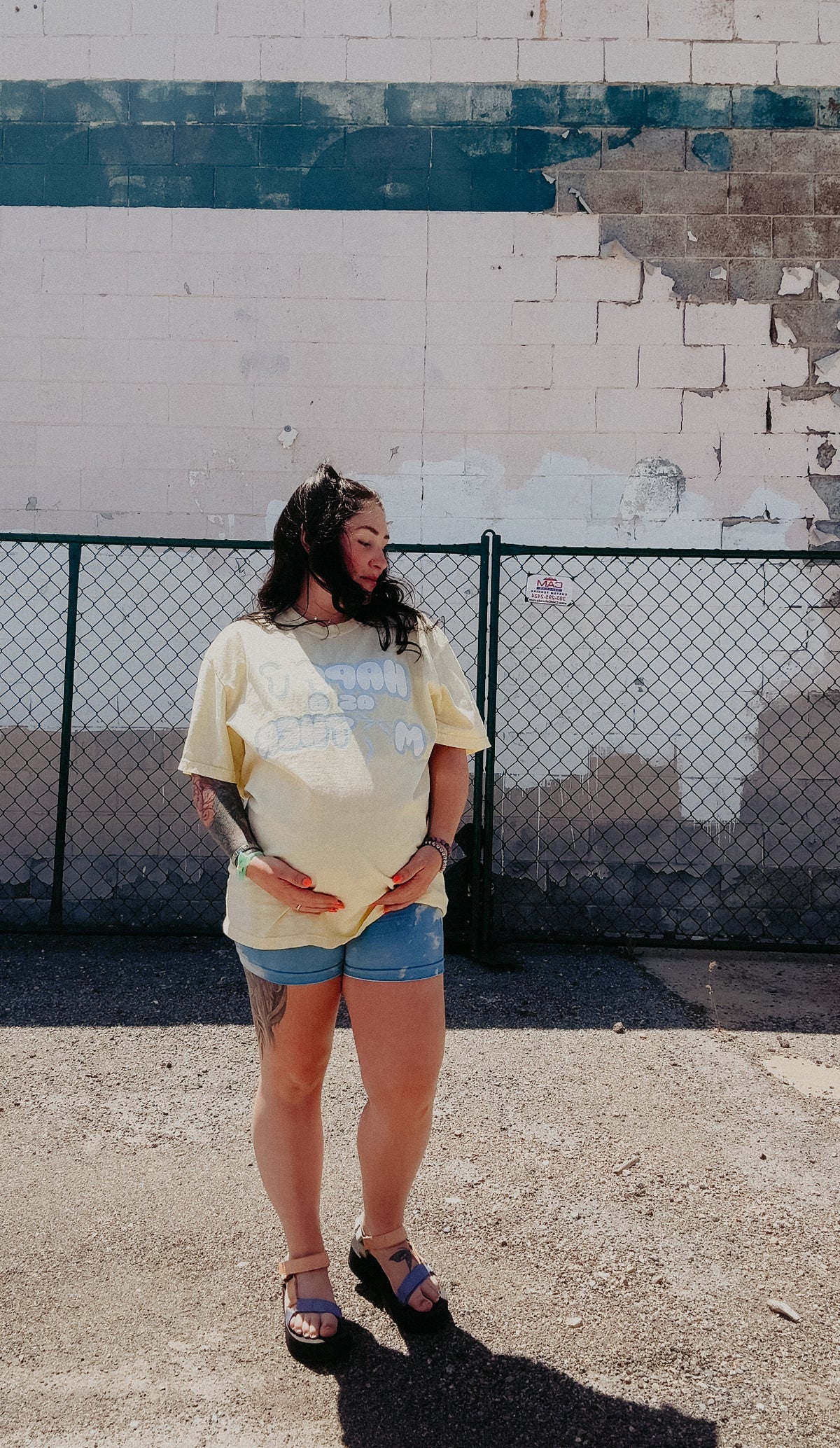 Pregnant mom wearing the LOVE WORLD Happy As A Mother Tee in Banana — oversized yellow garment-dyed tee with retro bubble letter graphic, styled with denim shorts and platform sandals