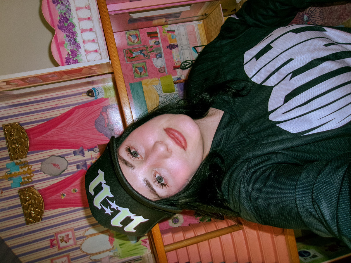 Woman wearing the LOVE WORLD 11:11 Trucker Hat in black with neon green gothic lettering — styled with MOTHER graphic hoodie, playful dollhouse backdrop