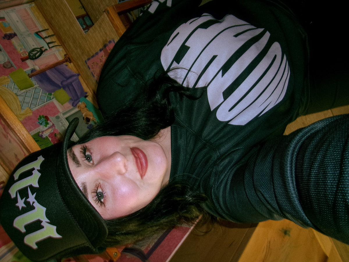 Close-up of woman wearing the LOVE WORLD 11:11 Trucker Hat — neon green gothic lettering on black, styled with MOTHER graphic hoodie