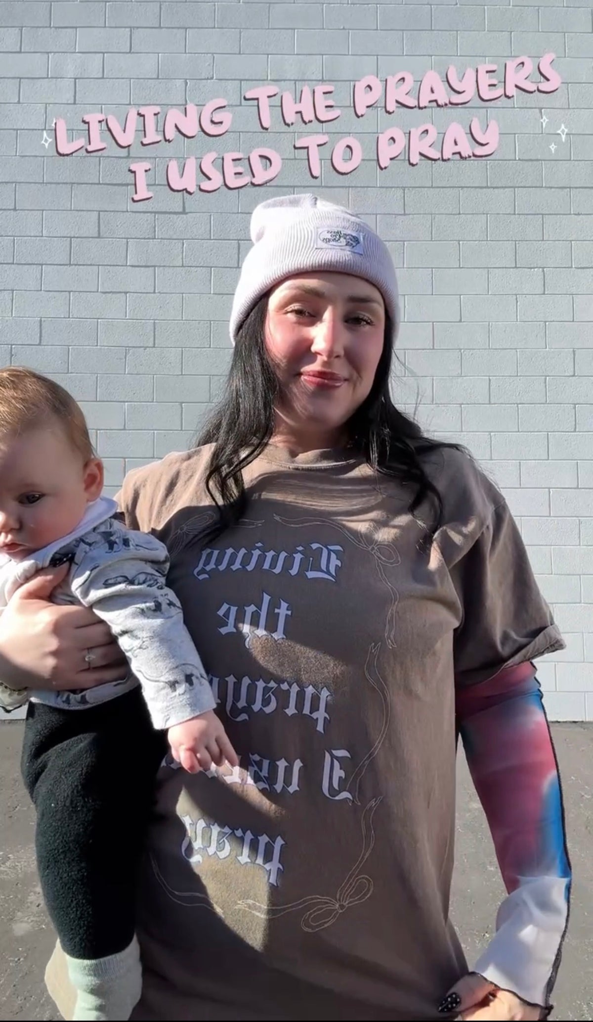 Model wearing LOVE WORLD Living the Prayers I Used to Pray Tee in Espresso — oversized Comfort Colors faith graphic tee, holding baby in front of brick wall with "Living the Prayers I Used to Pray" text