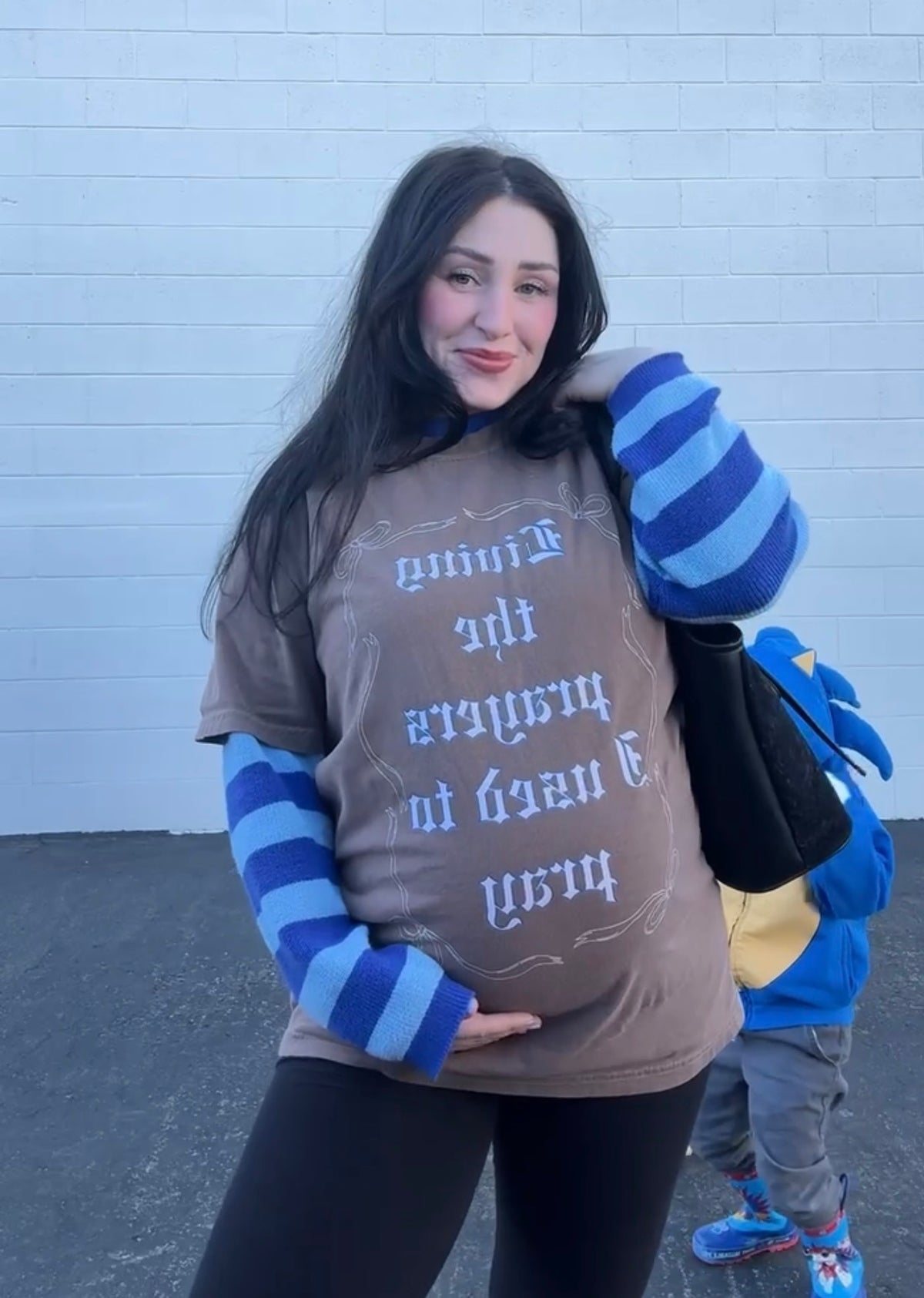 Model wearing LOVE WORLD Living the Prayers I Used to Pray Tee in Espresso — oversized Comfort Colors graphic tee, styled with striped long-sleeve underneath, holding baby outdoors