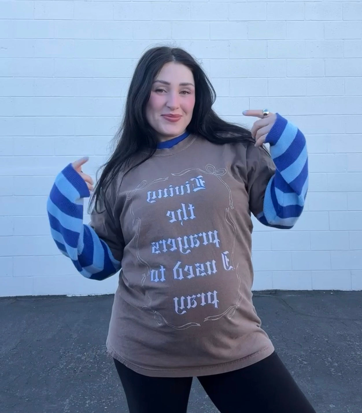 Model wearing LOVE WORLD Living the Prayers I Used to Pray Tee in Espresso — oversized Comfort Colors faith graphic tee, walking while holding baby, outdoor brick wall with "Living the Prayers I Used to Pray" text