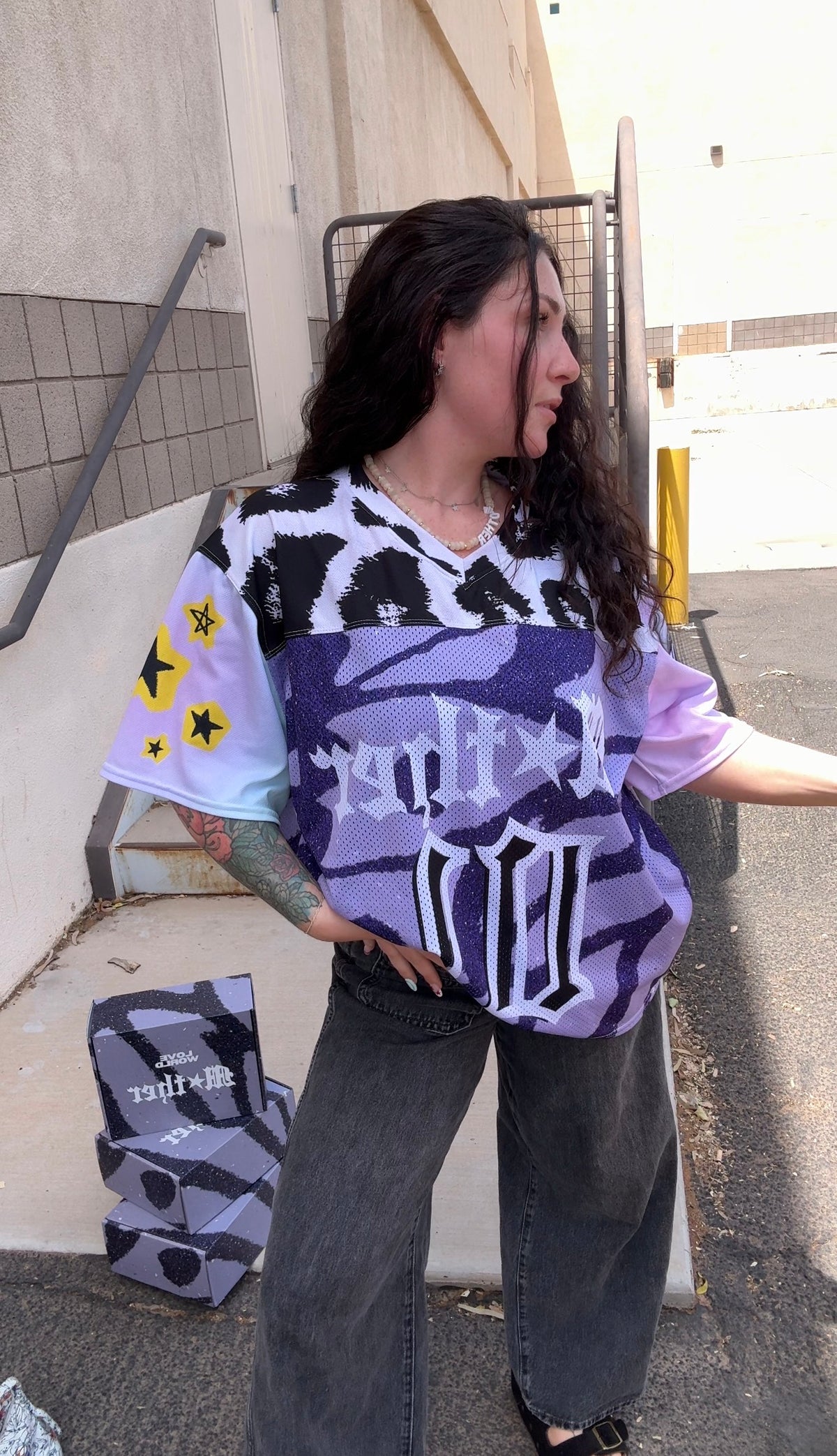 A woman wearing a purple, black, and white graphic t-shirt with gothic lettering and star details, paired with dark gray jeans, standing outdoors next to a building wall.mother jersey, leopard jersey, purple womens jersey, loveworldllc