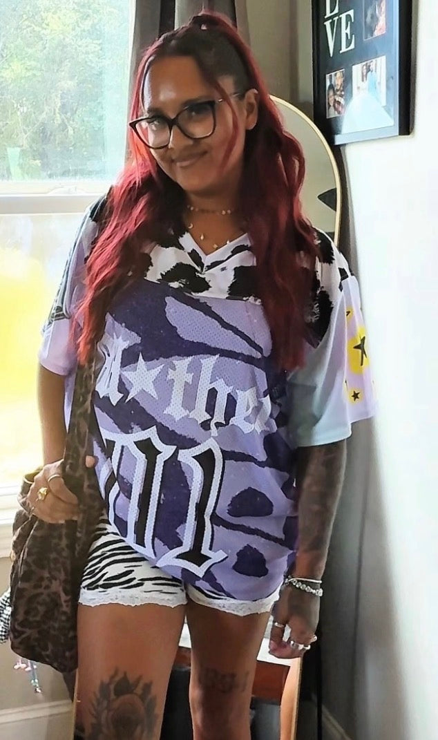 A woman with red hair and glasses wearing a purple and white graphic t-mother jersey with a zebra print sleeve, paired with white zebra-print shorts, standing indoors.