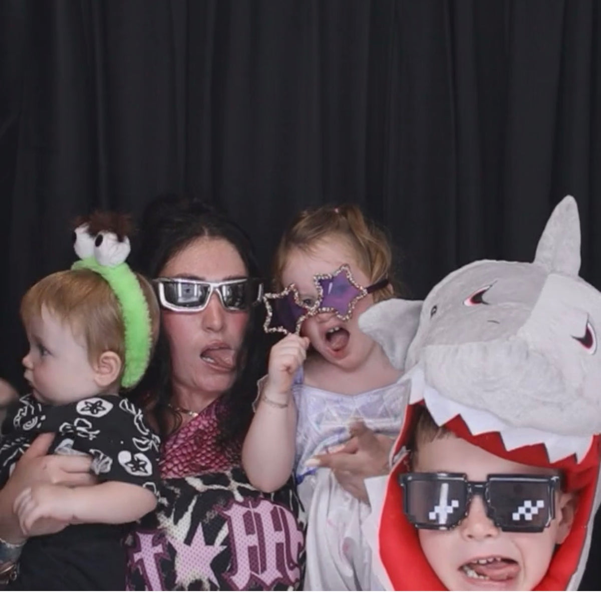 Three people posing together in a photo booth wearing funny costumes and accessories: a baby in a green headband with a small plush, a woman in silver 3D glasses, and a child in a gray shark costume with pixel sunglasses.