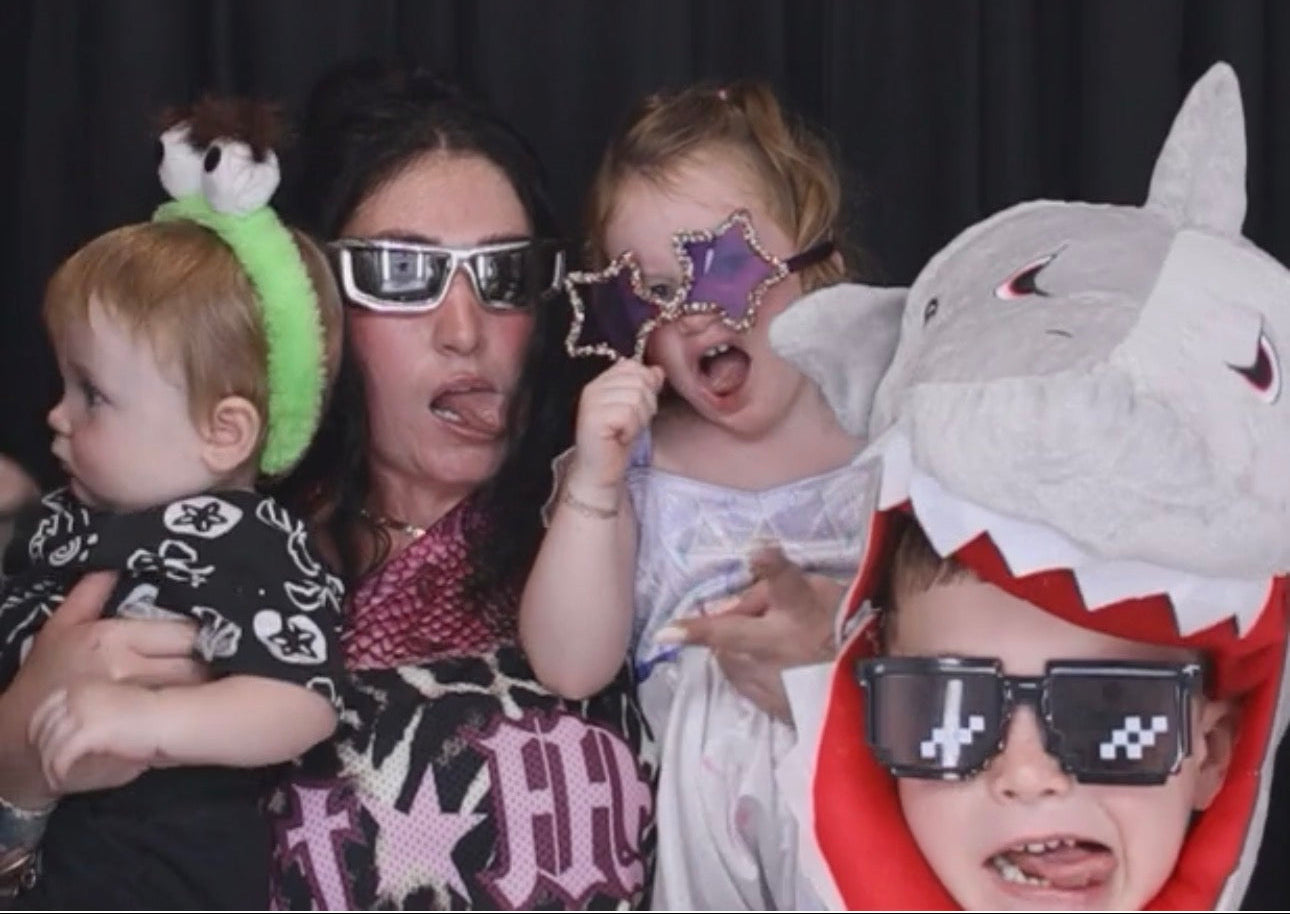 photo booth wearing funny costumes and accessories: a baby in a green headband with a small plush, a woman in silver 3D glasses, and a child in a gray shark costume with pixel sunglasses, taisha taylor loveworldllc clothing brand owner about 