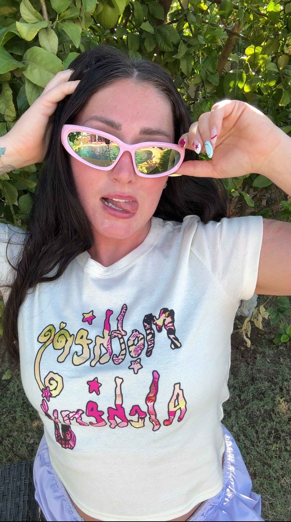 
Woman wearing the LOVE WORLD Mothers Alchemy cropped baby tee in natural/cream — pink and yellow retro graphic on fitted crop, styled with lavender shorts outdoors in front of a lemon tree