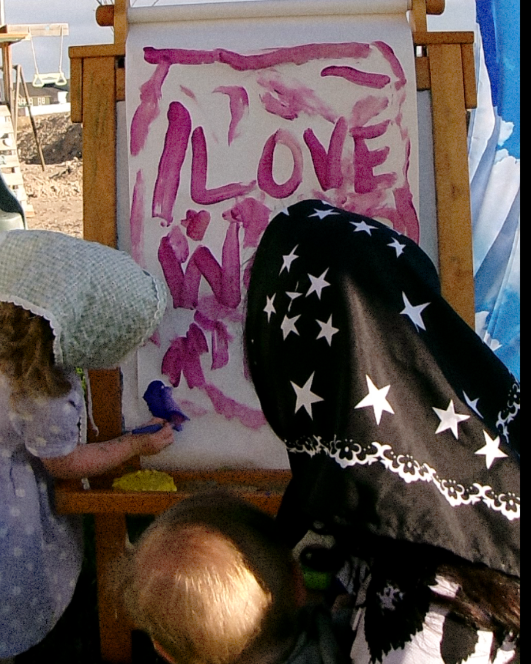 A child in a white bonnet paints on a canvas set up on an easel, with another person in a black and white star-patterned head covering partially blocking the view. The canvas features the word 'LOVE' painted in pink.womens graphic jersey, jersey for women, trendy top for women, womens clothing brand, dopamine dressing,dopamine clothing brand, t-shirt with a colorful graphic reading mother with snakeskin and leopard design, loveworldllc.com, stars and leopard jersey for women, pinterest ootd,star bandana