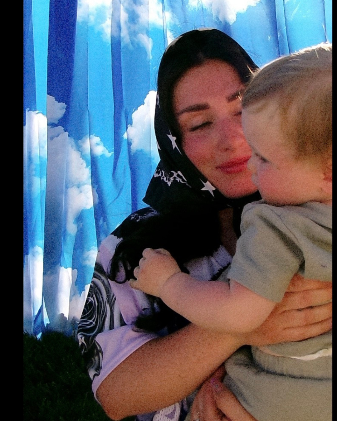 A woman wearing a black headscarf with white star patterns holds a baby against a blue sky and clouds printed backdrop.womens graphic jersey, jersey for women, trendy top for women, womens clothing brand, dopamine dressing,dopamine clothing brand, t-shirt with a colorful graphic reading mother with snakeskin and leopard design, loveworldllc.com, stars and leopard jersey for women, pinterest ootd,star bandana